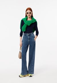 Cardigan a coste blu navy, sciarpa in maglia verde, jeans in denim a gamba larga a vita alta e sneakers con dettagli neon. Borsa beige.