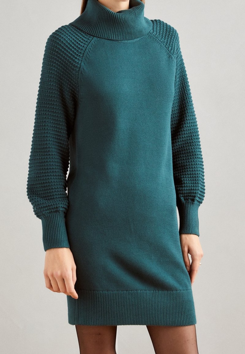 Robe tricotée turquoise avec col roulé, manches bouffantes texturées et poignet côtelé. Corps lisse, avec des accents sur les épaules et les manches.