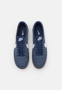 Nike sportbačiai su tamsiai mėlynu zomšiniu paviršiumi, baltais logotipais, plokščiomis tamsiai mėlynomis raištelėmis, paminkštintu apykaklės kraštu ir rudą guminę padu.
