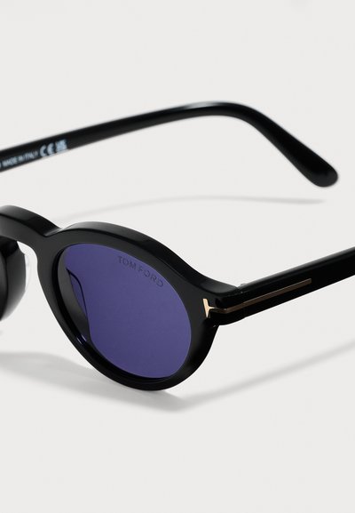 Lunettes de soleil rondes noires avec des verres bleu foncé et des accents dorés sur les branches, présentées sur un fond blanc.