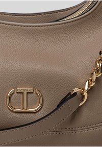 Taupe leren handtassectie met gouden Twinset-logo en kettingdetail op het handvat, waarbij de textuur en stiksels zichtbaar zijn.