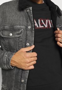 Chaqueta de mezclilla negra con cuello de lana, botones a presión y bolsillos en el pecho, que se lleva sobre un sudadera negra con un diseño de texto en rojo y blanco.