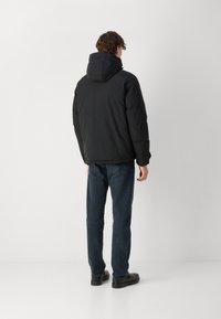 Musta toppatakki hupulla, sileä pinta ja joustavat hihansuut, yllään tummansiniset farkut ja mustat kengät, kuvattuna takaa.