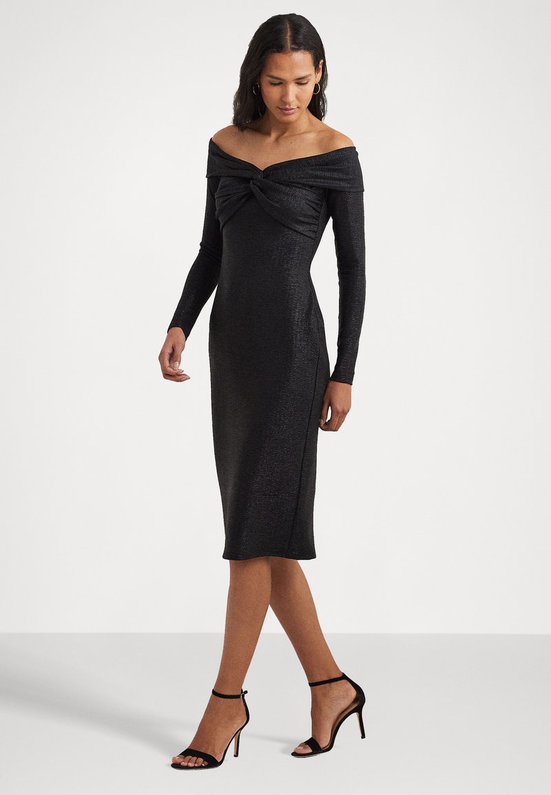 Abito nero off-shoulder con maniche lunghe, dettaglio annodato sul davanti, tessuto liscio e taglio fino al ginocchio, abbinato a tacchi neri con cinturino alla caviglia.