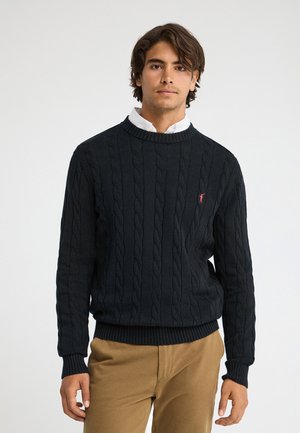 Joven con cabello castaño de longitud media que lleva un suéter de punto trenzado negro sobre una camisa blanca y pantalones color tostado, de pie frente a un fondo liso.