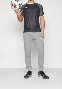 Conjunto atlético que incluye una camiseta de patrón negro y gris, pantalones de chándal grises y zapatillas deportivas negras con detalles blancos, sosteniendo un balón de fútbol.