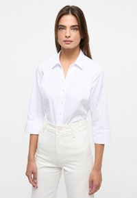 Camicia bianca con colletto e maniche a tre quarti, caratterizzata da un tessuto texturizzato. Abbinata a pantaloni bianchi a vita alta.