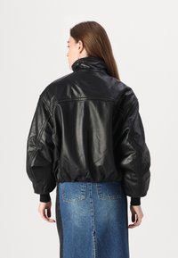 Veste bomber en cuir noir avec un col haut et une coupe oversized, présentant des accents cousus et des poignets côtelés, associée à une longue jupe en denim.