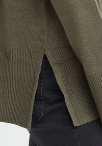 Pull en maille vert olive avec une fente latérale dévoilant un jean en denim foncé en dessous.