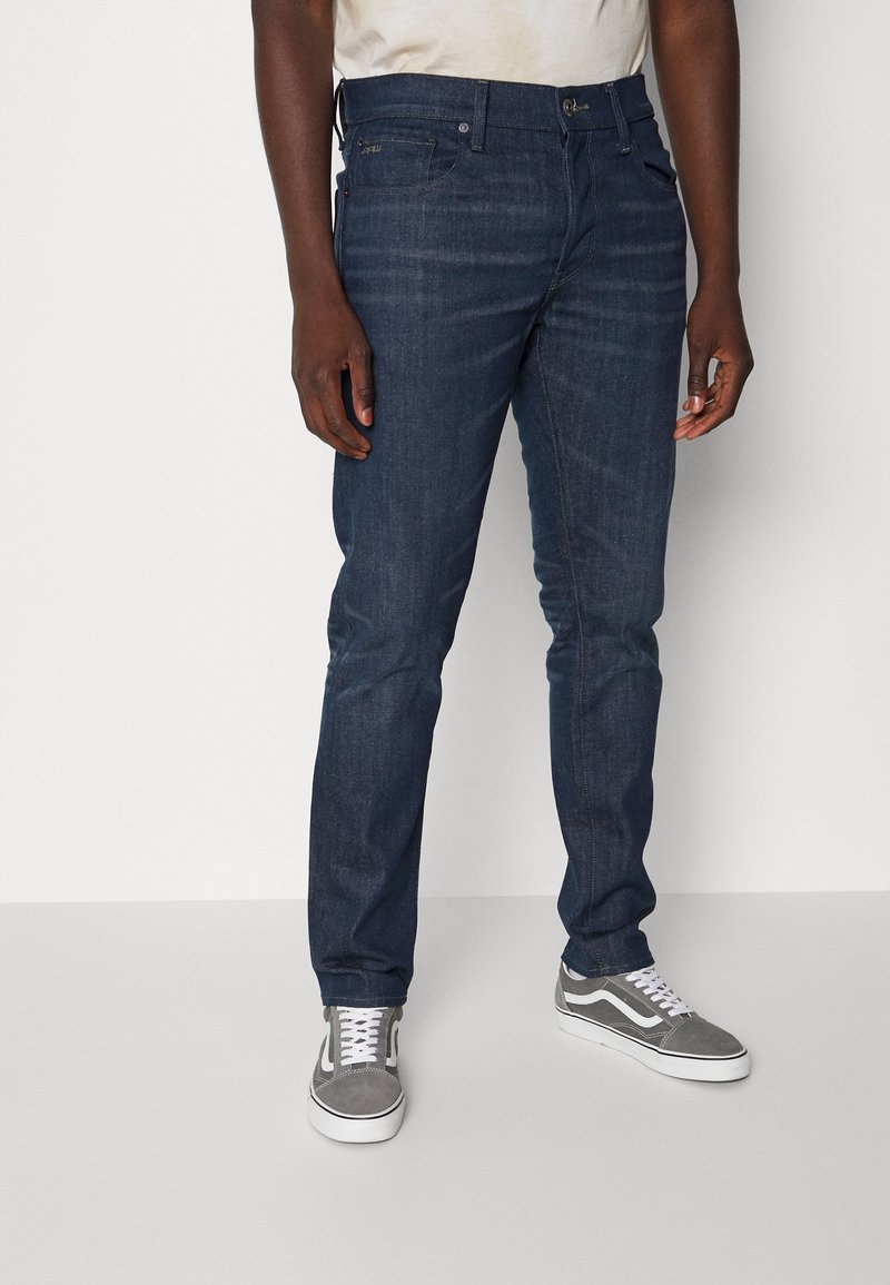 Jean en denim bleu foncé à coupe slim, avec un design classique à cinq poches, des décolorations subtiles et des détails de couture. Associé à des baskets grises.