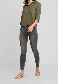 Mujer vistiendo una blusa abotonada verde oliva, jeans pitillo gris oscuro acanalados y sandalias beige de punta abierta con tiras de estampado animal en negro y marrón.