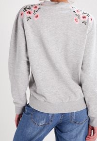 Felpa grigia con ricami floreali rosa sul collo posteriore, maniche a coste e orlo, abbinata a jeans in denim blu.
