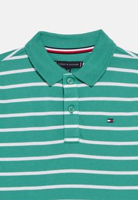 Πράσινη και λευκή ριγέ μπλούζα πόλο με κλασικό γιακά, τρία κουμπιά και ένα μικρό κεντημένο λογότυπο σε κόκκινο, λευκό και ναυτικό μπλε.