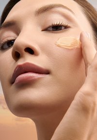 Gros plan sur une femme appliquant une crème beige clair sur sa joue. La peau est lisse avec un ton neutre ; un maquillage naturel met en valeur les traits.