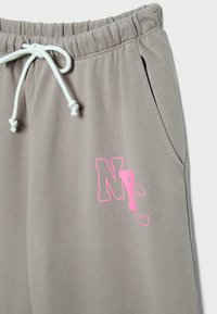 Hellbraune Sweatpants mit elastischem Bund, weißem Kordelzug, zu einer Schleife gebunden, seitlicher Tasche und rosa "NYC"-Grafik auf dem linken Oberschenkel.