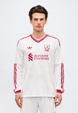 Jeune homme portant un maillot blanc à manches longues du club de football de Liverpool avec des bandes rouges et le logo Standard Chartered, debout devant un fond uni.