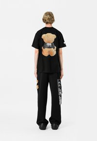 Camisa negra de manga corta con un diseño de un osito de peluche difuso en la parte de atrás, combinada con pantalones negros y zapatos negros. Look sencillo y casual.