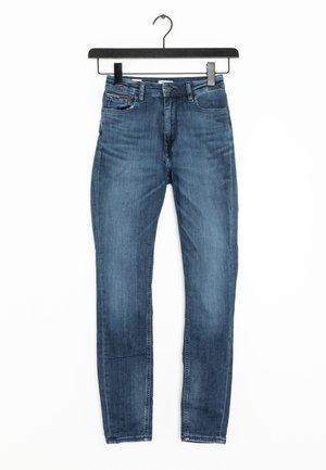 Niebieskie dopasowane dżinsy z pięcioma kieszeniami, zapięciem na guzik i szlufkami na pasek, wiszące na czarnym wieszaku na białym tle.