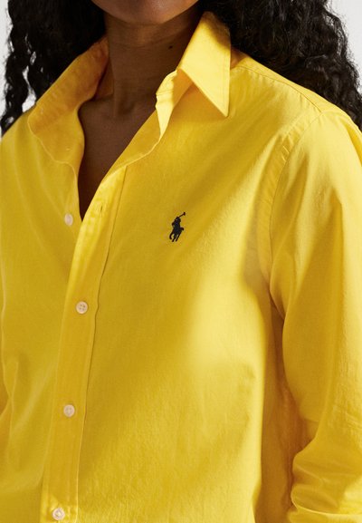 Asmuo dėvi ryškiai geltoną marškinius su sagutėmis, ant krūtinės yra mažas juodas polo žaidėjo logotipas, matomi garbanoti plaukai.