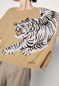 Pull en maille beige avec un grand motif de tigre blanc aux rayures noires, ourlet côtelé, et le nom de la marque "TWINSET" brodé en bas.