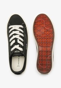 Svarte lerretssko med hvite lisser, med rund tå, gummisåle med et teksturert rødt mønster, og Lacoste-logo på siden.
