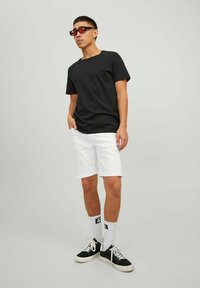 Camiseta negra de mangas cortas, pantalones cortos de mezclilla blancos, zapatillas negras con detalles blancos y gafas de sol rojas. Fondo liso.