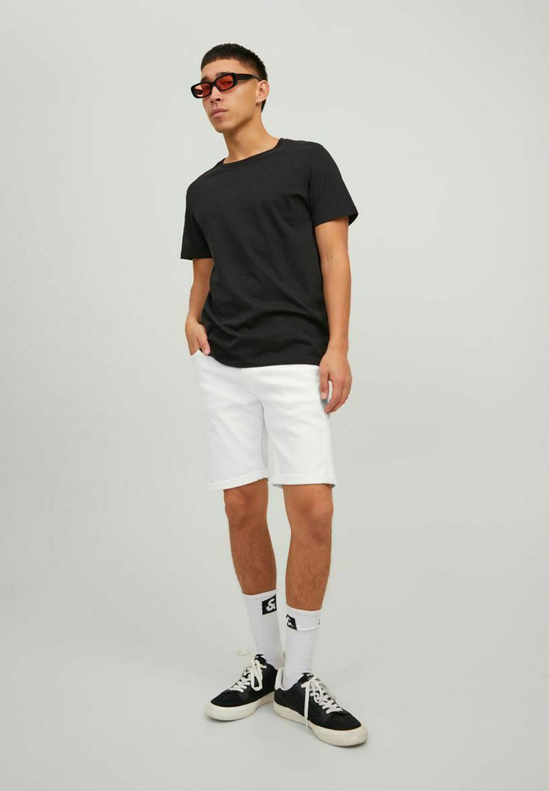 Camiseta negra de mangas cortas, pantalones cortos de mezclilla blancos, zapatillas negras con detalles blancos y gafas de sol rojas. Fondo liso.