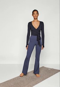 Top în stil wrap de culoare marină, cu mâneci lungi și decolteu adânc în V, asortat cu pantaloni de yoga flared, toată ținuta fiind realizată dintr-un material moale și elastic.
