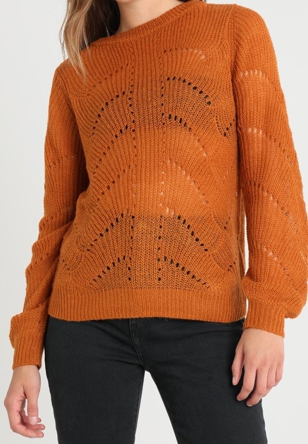 Femme portant un pull en tricot orange brûlé texturé avec des trous à motifs et des manches longues, associé à un pantalon noir.