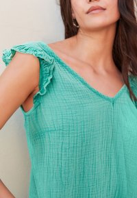 Lichtgroene, getextureerde top met een diepe V-hals en gerimpelde schouderaccenten. De stof lijkt zacht en licht gekreukt.