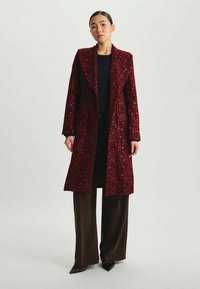 Manteau long en sequins bordeaux avec un col incrusté, un tissu texturé et un design ouvert. Stylé sur un pantalon marron à jambes larges.