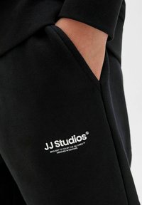 Svarta joggers med fickor som har en mjuk textur och vit logotext "JJ Studios" nära fickan. Designen är enkel och funktionell.