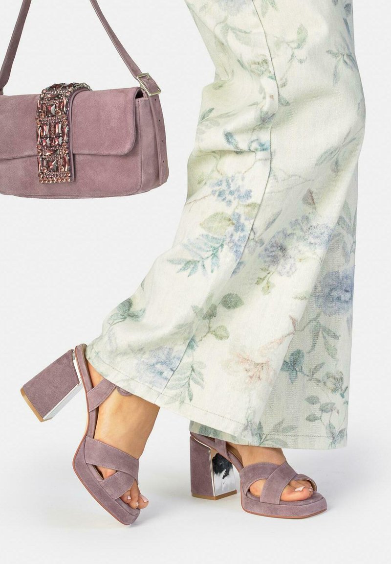 Kobieta ubrana w kwieciste spodnie z szerokimi nogawkami, sandały na słupku zamszowe w kolorze mauve oraz trzymająca dopasowaną ozdobioną torebkę na ramię w kolorze mauve.