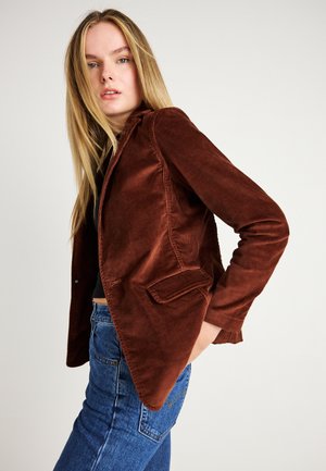 Brun fløjl blazer med en skræddersyet pasform, notch-kraver, enkelt knaplukning og to forlommer, parret med blå jeans.
