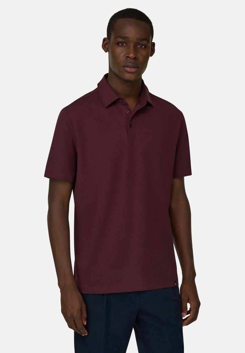 Muž oblečený do tmavě červené polokošile s krátkými rukávy a tmavě modrých kalhot, stojící před jednobarevným světlým pozadím.