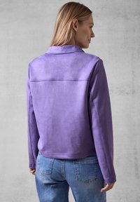 Veste en suede violet avec un col et des manches longues, présentant une texture lisse et des détails de couture simples. Portée avec un jean bleu.