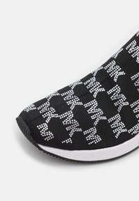 Schwarzer Sneaker mit einer weißen gepunkteten Muster, das abstrakten Text bildet, weißer Zwischensohle und strukturiertem schwarzen Außensohle vor einem hellen Hintergrund.