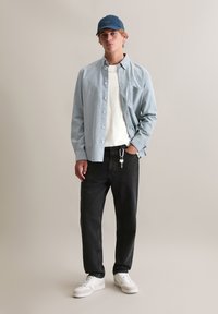 Jasnoniebieska koszula na guziki noszona na białej koszulce, w połączeniu z czarnymi dżinsami i białymi trampkami. Dodatki obejmują niebieską czapkę i breloczek.