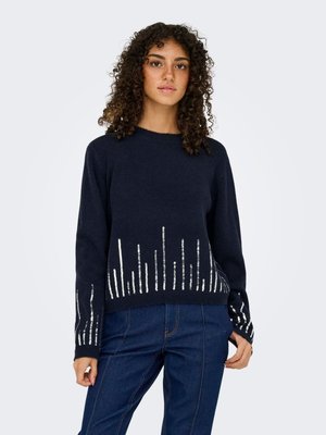 Jonge vrouw met krullend haar die een donker marineblauwe trui draagt met verticale zilveren paillettenstrepen en een high-waisted blauwe spijkerbroek, staand tegen een witte achtergrond.