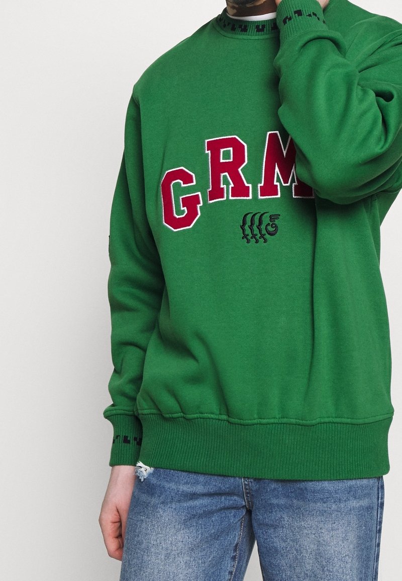 Felpa verde con colletto a girocollo, decorata con la scritta ricamata in rosso "GRM" e un design grafico. Polsini e orlo a costine, morbida al tatto.
