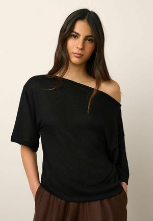 Fiatal nő hosszú sötét hajjal, fekete vállat szabadon hagyó felsőben és barna nadrágban, zsebébe tett kézzel áll egy egyszínű háttér előtt.