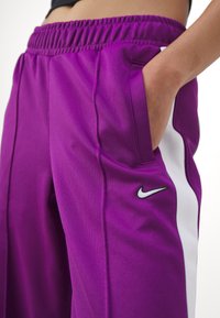 Purple athletic pants with white side stripes, elastic waistband, and side pockets. Features a small black Nike logo on the lower leg.