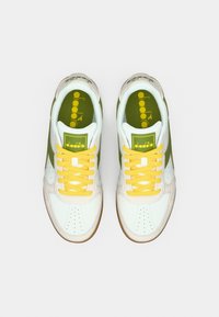 Λευκά δερμάτινα αθλητικά παπούτσια με πράσινες λεπτομέρειες, κίτρινα κορδόνια και διάτρητη μύτη. Περιλαμβάνει υφή από μπεζ σουέντ και λογότυπο στη γλώσσα.