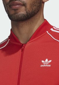 Red zip-up jacket with a high collar, featuring white stripes on shoulders and a small white Adidas logo on the chest. Smooth fabric texture.