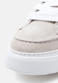 Δερμάτινο αθλητικό παπούτσι σε ανοιχτό μπεζ χρώμα με λευκή καουτσούκ σόλα, διαθέτει χοντρές υφασμάτινες κορδόνια και ραμμένες λεπτομέρειες κατά μήκος των άκρων.