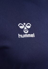 Tissu bleu marine avec un logo "hummel" blanc représentant une abeille stylisée. Texture lisse, design minimaliste et logo placé au centre.