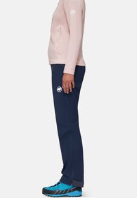 Jasnoróżowa bluzka z długim rękawem, z kieszonką z przodu i logo, w połączeniu z granatowymi spodniami z zamkami i logo, noszona z niebieskimi butami turystycznymi.