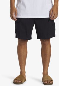 Black cargo shorts with multiple pockets, made from textured fabric, paired with brown slide sandals featuring a wide strap design.