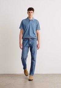 Cămașă polo albastră deschis cu mâneci scurte, asociată cu blugi clasici albastre și cizme maro. Fundalul este neutru, cu un design simplu.