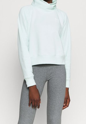 Person trägt hellblauen Kapuzenpullover und graue Leggings, steht mit locker an den Seiten hängenden Armen vor einem einfarbigen Hintergrund.